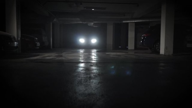 Dark underground parking lot concrete floor car headlights backlight dolly in gliding low angle shot