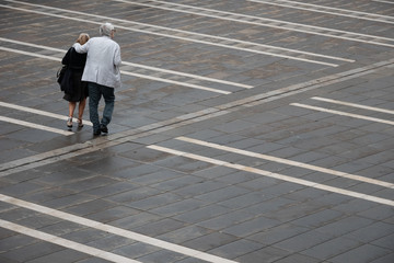elderly, well dresed couple is crossing a paved square structured by white lines
