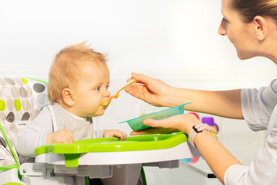 Mother Feed Little Toddler Baby Boy From Plastic Spoon In High Chair View From Side