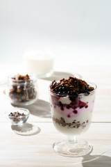 Yogurt parfait with cocoa granola, frozen blueberries and yogurt in bokeh. White wooden table, hard light, high resolution