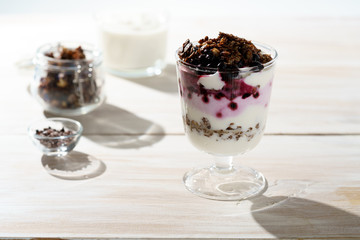 Yogurt parfait with cocoa granola, frozen blueberries and yogurt in bokeh. White wooden table, hard light, high resolution