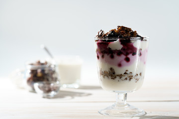 Yogurt parfait with cocoa granola, frozen blueberries and yogurt in bokeh. White wooden table, hard light, high resolution