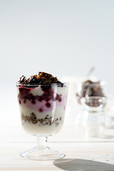 Yogurt parfait with cocoa granola, frozen blueberries and yogurt in bokeh. White wooden table, hard light, high resolution