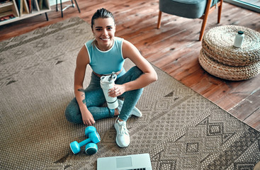 A sporty woman in sportswear is sitting on the floor with dumbbells and a protein shake or a bottle...