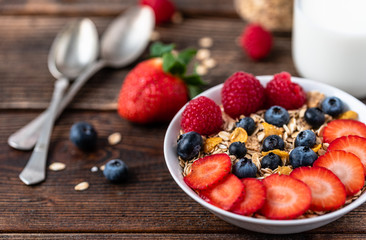 Granola with blueberries and raspberries in white bowl in dark wooden desk.