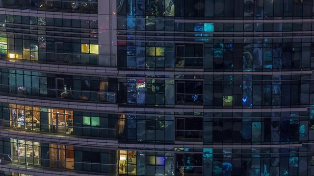 Rows of glowing windows with people in the interior of apartment building at night. Modern skyscraper with glass surface. Concept for business and modern life. Zoom out