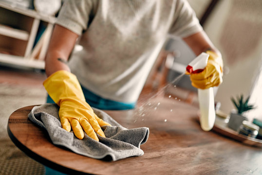 Cropped image of a beautiful young woman makes cleaning the house. Girl rubs dust. Woman in protective gloves is smiling and wiping dust using a spray and a duster while cleaning her house.