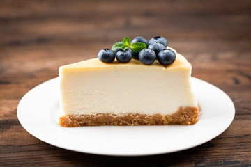 Plain New York Cheesecake Slice Topped With Blueberries And Mint Leaf On White Plate Wooden Table Background. Tasty Slice Of Creamy Cake - obrazy, fototapety, plakaty