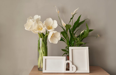 Bouquet of fresh white tulips and empty photo frames with copy space on shelf in front of wall. Mock up with Frame and white Tulips. Mother's Day, Women's Day, March 8, spring greeting card template.