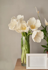 Bouquet of fresh white tulips and empty photo frames with copy space on shelf in front of wall. Mock up with Frame and white Tulips. Mother's Day, Women's Day, March 8, spring greeting card template.