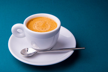 White espresso cup and saucer with brown coffee on a teal blue surface with beans and spoon.