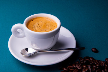 White espresso cup and saucer with brown coffee on a teal blue surface with beans and spoon.