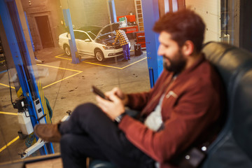 man looking at his smartphone with mechanic man repairing his car in workshop