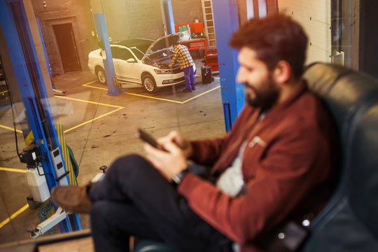 man looking at his smartphone with mechanic man repairing his car in workshop