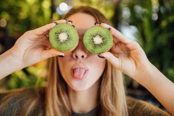 Happy young and blonde woman holding kiwi in front of eyes
