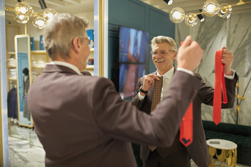 A happy millionaire in a boutique trying on a tie near a large mirror.