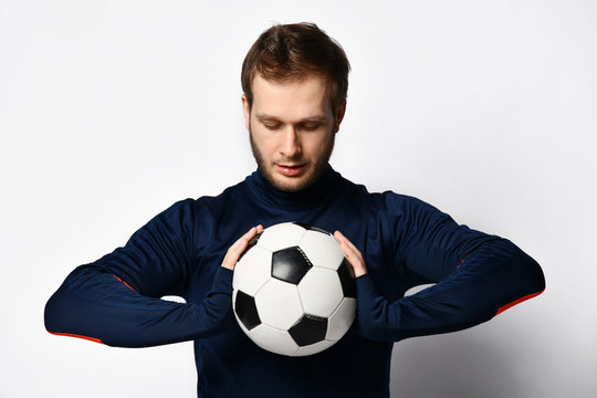 Skillful soccer player in blue tracksuit is smiling and holding a ball, posing isolated on white studio background. Close up