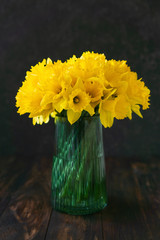 Yellow narcissus flower bouquet in a vase on wooden table, dark background, natural sunny light, copy space. Greeting card for Easter, Mothers, Woman Day, birthday, spring concept.