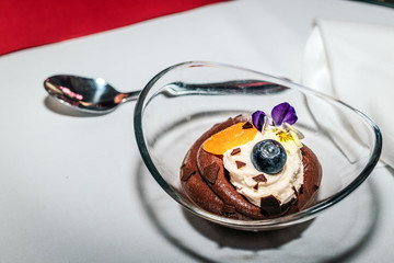 Mousse au Chocolat in ovaler Glasschüssel mit Heidelbeere, Mandarine und  Stiefmütterchen als Dekoration sowie Löffel im Hintergrund