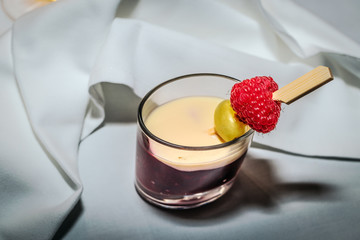 Rote Grütze in ovalem Dessertglas mit Vanillesoße, Weinbeere und Himbeere am Spieß auf weißem Tafeltuch