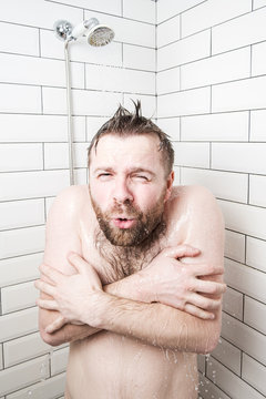 Man, with a funny expression on his face, feels shocked at taking a cold shower, he froze and tries to cover his body with hands.