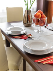 Tableware and a big bowl of fruit on a dining table in daylight, angled view.