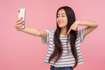 Naklejka premium Portrait of charming asian girl with brunette hair taking selfie on mobile phone, communicating on video call and pointing down, gesturing subscribe. indoor studio shot isolated on pink background