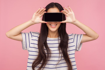 Anonymous mobile user. Portrait of girl with long brunette hair in striped t-shirt hiding eyes with cell phone and smiling, unknown internet customer. indoor studio shot isolated on pink background