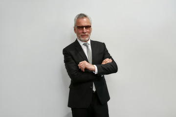 Looking perfect. Portrait of confident mature businessman in eyeglasses keeping arms crossed and looking at camera while standing against grey background