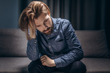 Mature man in casual clothing leaning with elbows on knees and sitting on couch with sad expression. Bearded person feeling depressed at home.