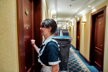Let us clean. Smiling hotel maid in uniform knocking on the door for room service while standing with the chambermaid trolley in the hall.