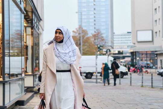 Young stylish Arabic woman in hijab dreamily walking around street with shopping bags