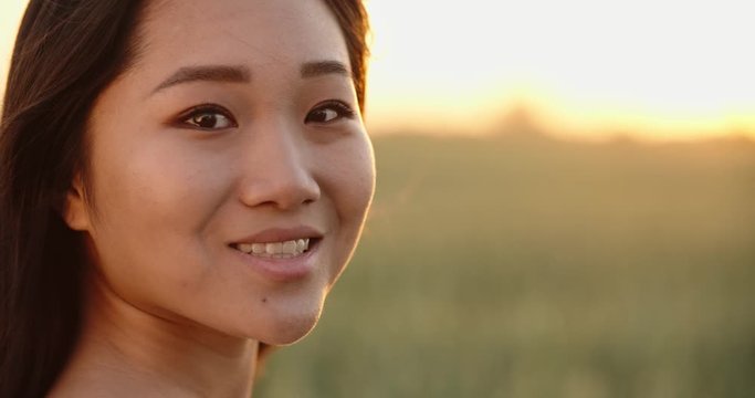 Portrait shot of panasian girl standing in golden wheat field during bright sunset, with wind blowing ito her hair - close up 4k footage