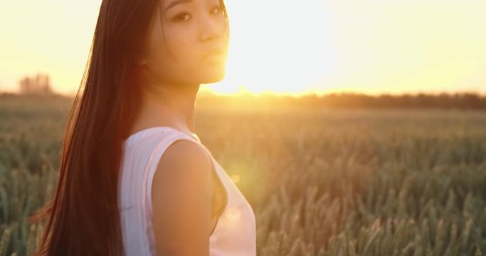 Portrait shot of panasian girl standing in golden wheat field during bright sunset, with wind blowing ito her hair - close up 4k footage