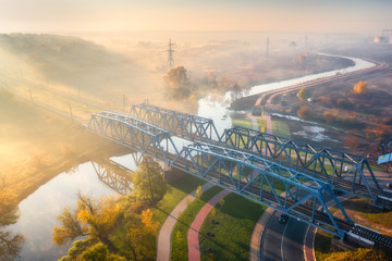 Aerial view of beautiful railroad bridge and river in fog at sunrise in autumn. Industrial landscape with railway station,  gold sun rays, road, mist, trees, orange sky in foggy morning. Top view