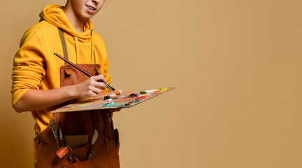 Young teen boy painter in yellow hoodie and jumpsuit standing and painting on easel over yellow background