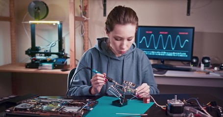 A woman engineer solders circuits sitting at a table. Microchip production factory. Technological process. Assembling the PCB board. Girl repairing electronic device on the circuit board. - Powered by Adobe