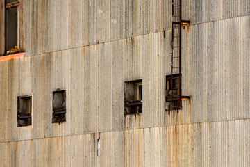 A large exterior concrete wall of an old industrial building. There are four small windows and an iron ladder on the wall. The textured building has rust stains under the latter and windows. 