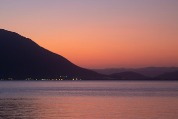 Obraz premium Sunset with orange and magenta tones on the sky reflecting in the sea with mountains and some lights at the horizon seen from Florianópolis