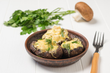 Baked mushrooms in a clay bowl and a fork on a white wooden table.