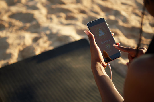 Close-up image of sportswoman standing on sandy beach and starting yoga session on smartphone