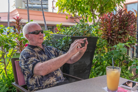 A senior man takes a photo with his phone's camera at a restaurant surrounded by a garden.