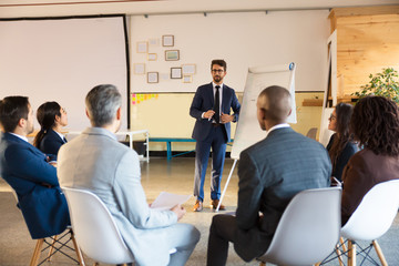 Smiling business trainer working with employees. Group of workers sitting on chairs and listening...