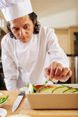 Mature Vietnamese chef garnishing baked chicken in tray with fresh dill