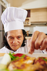 Smiling mature man in chef hat garnishing chicken he baked in tray with parsley