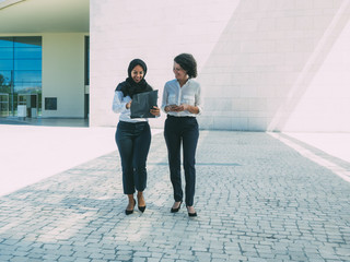 Happy confident female business partners discussing contract outside. Multiethnic women in office suits and hijab walking outside and carrying folder with documents. Partners concept