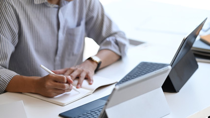Cropped image of smart man working as Project administrator writing on notebook while sitting in front computer tablet with keyboard case with the modern office as background