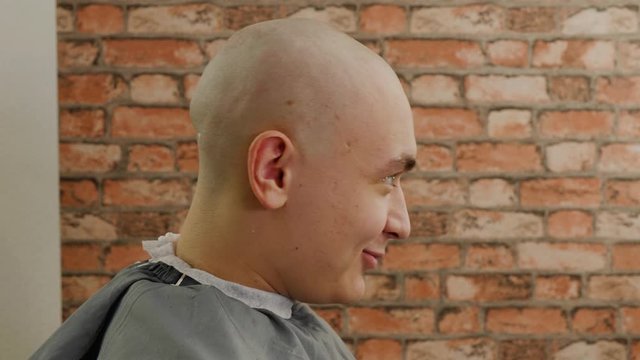 Bald man with shaved head sitting in barber chair in male salon. Profile view bald man after shave talking in barbershop. Portrait young man in hairdressing salon