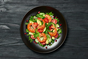 Shrimps salad with cherry tomatoes, cucumeber, avocado, lettuce and pomegranate on dish. Healthy seafood concept. Tasty grilled prawn shrimp and mix vegetable salad on black wood, top view.