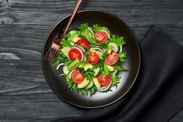 Fresh salad with cherry tomatoes, avocado,cucumeber and lettuce on dish. Healthy vegetarian meal concept. Tasty mixed leaves, tomatoes and mix vegetable salad on black wood, top view.